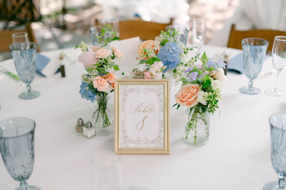 Elegant wedding reception table settings with blue glassware, peach flowers, and gold framed table numbers.