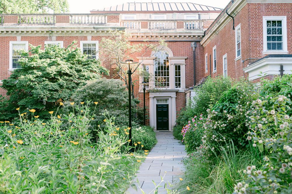 A serene garden pathway lined with lush greenery and brick buildings leads to an arched doorway.