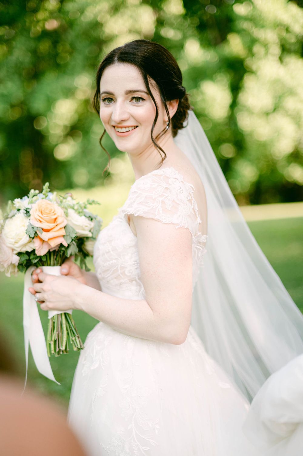 Bride holding peach and white floral bouquet in sunlit garden setting.