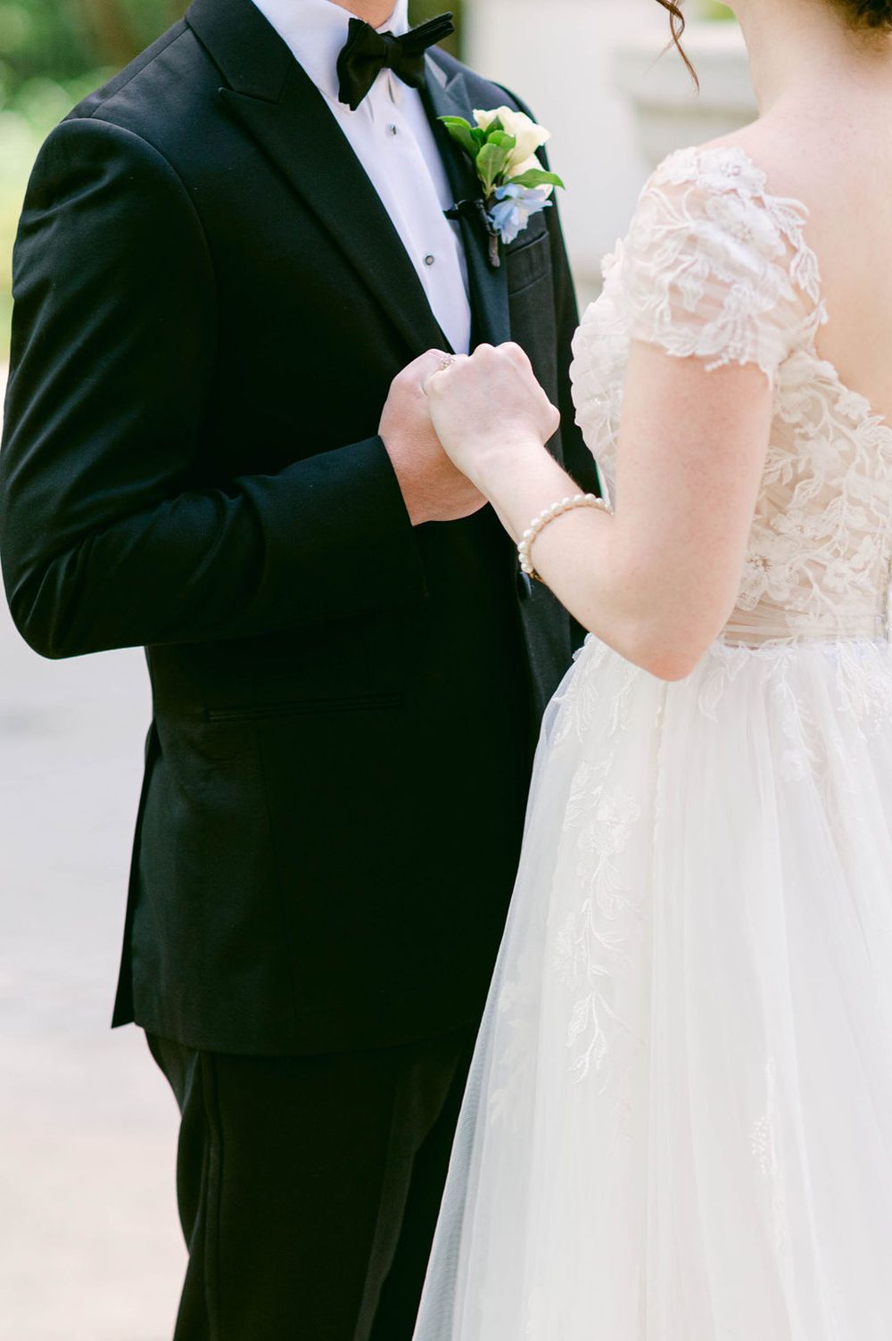 Close-up of bride and groom holding hands at wedding ceremony showing elegant white dress and black tuxedo details.
