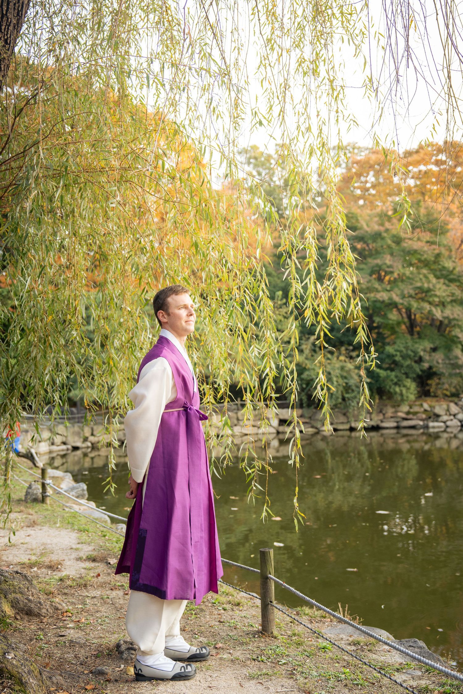 A figure in a purple coat stands near a tranquil pond surrounded by autumn willow trees in a serene park setting.