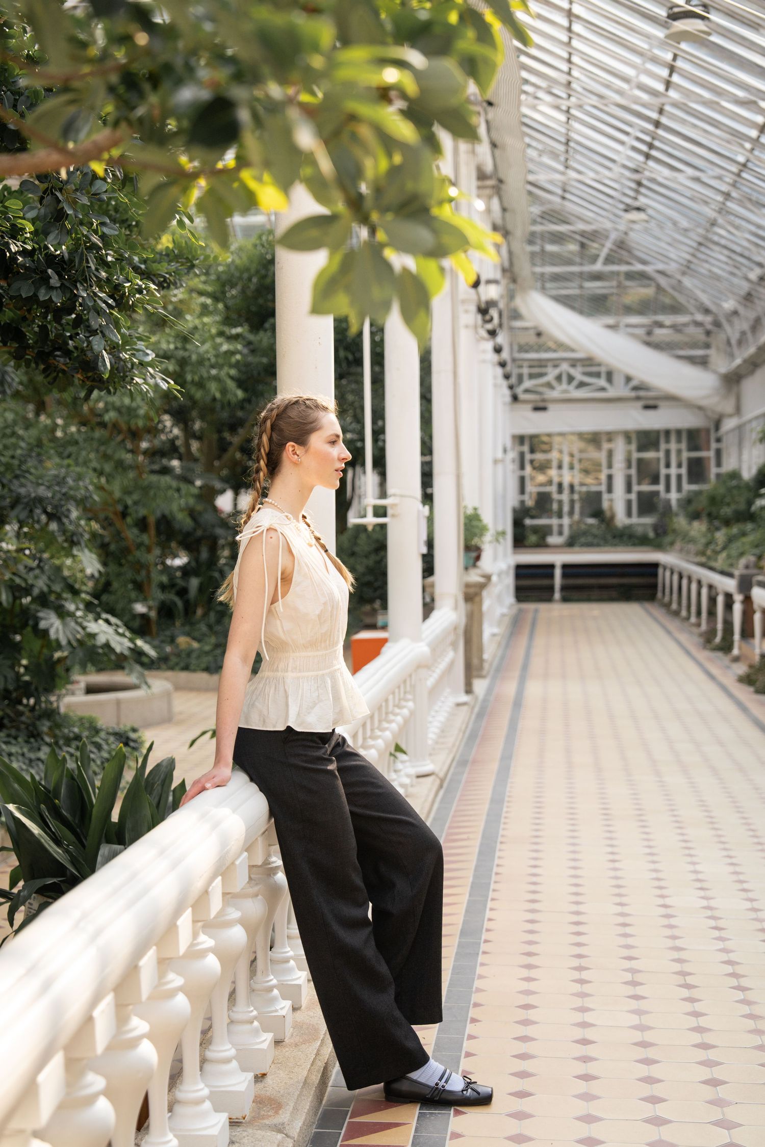 A person in vintage-style clothing leans against an ornate white balustrade in a sunlit greenhouse conservatory.