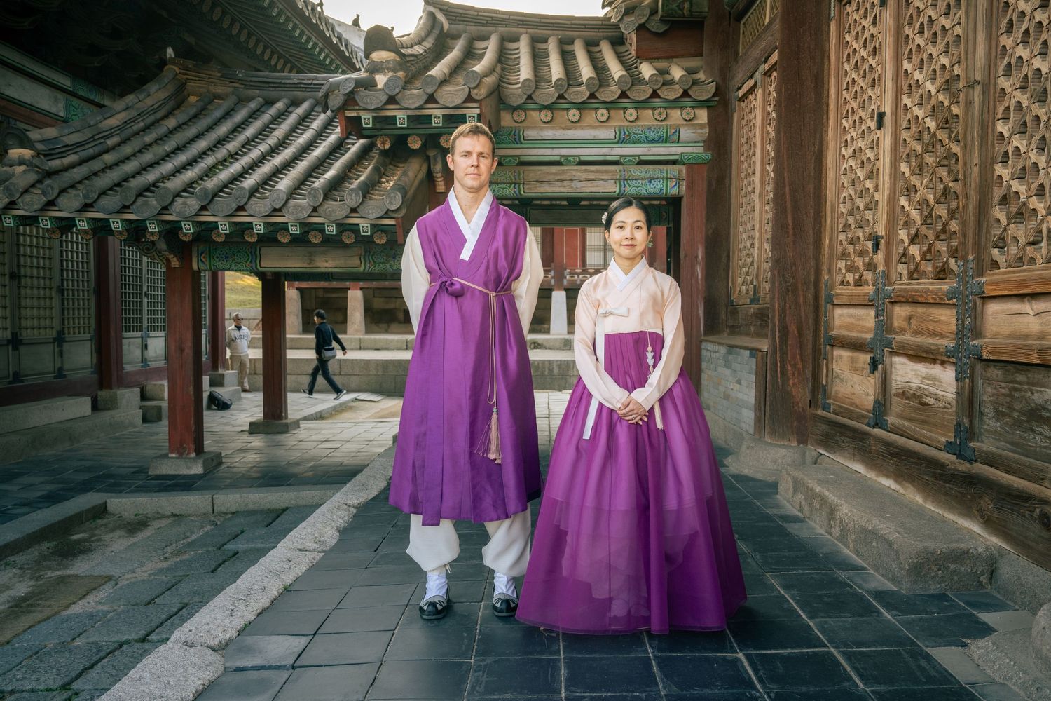 Traditional Korean architecture with ornate wooden buildings and walkways decorated in vibrant purple and white hanbok attire.