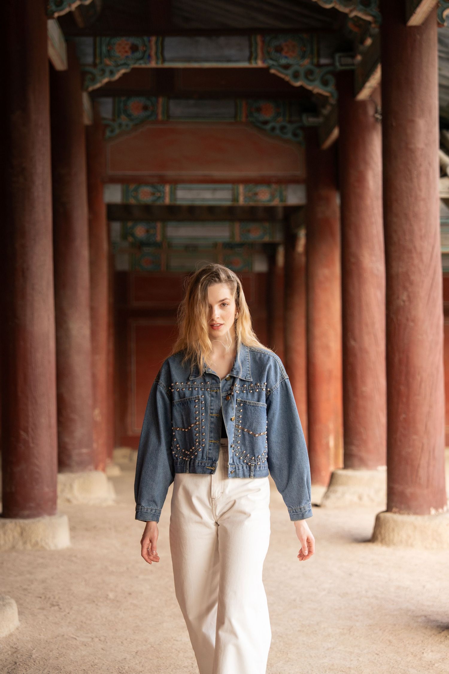 Person in denim jacket and white pants walks through traditional Asian temple corridor with red wooden pillars.