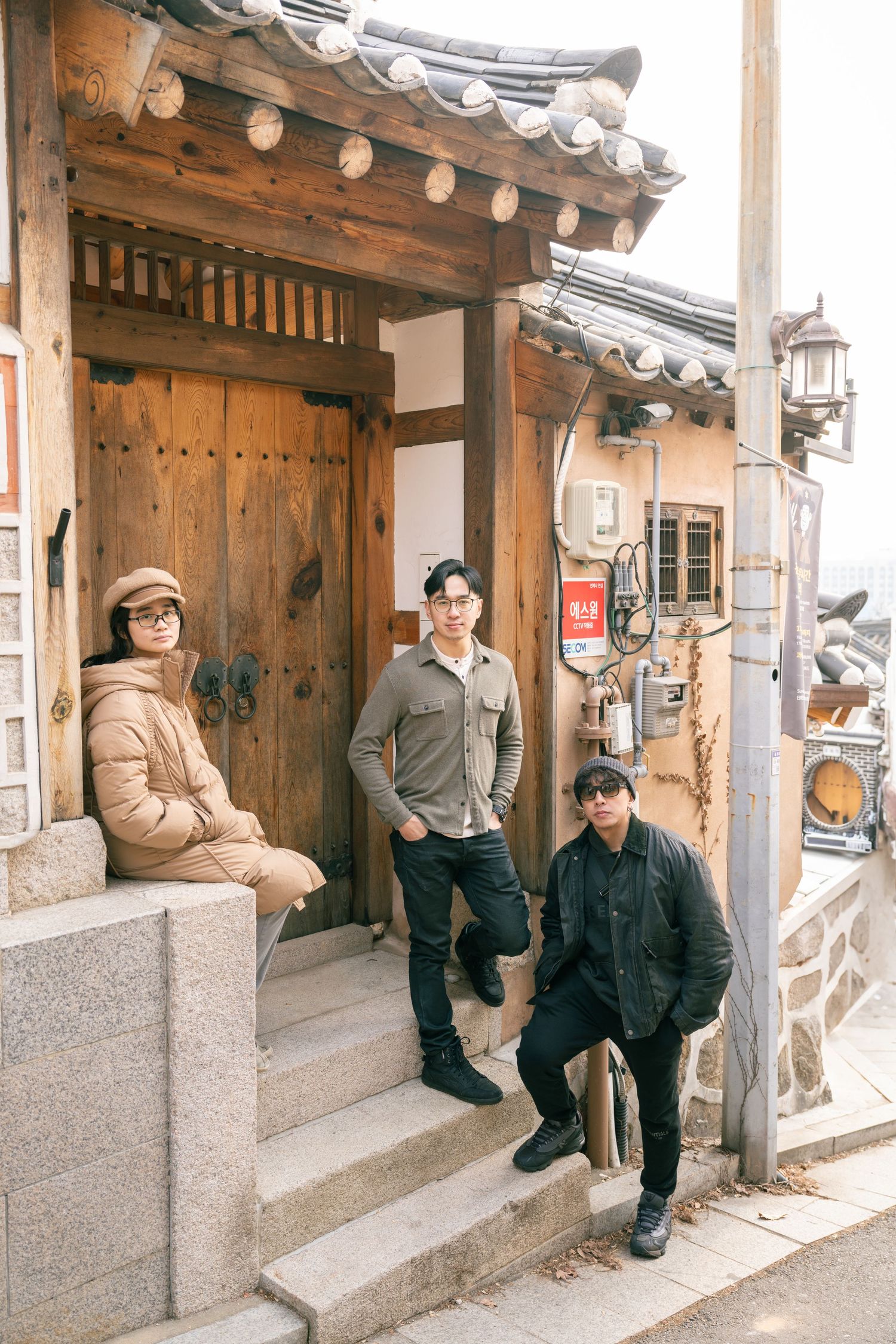 People sit along a traditional Korean hanok house wall with tiled roof and wooden architecture in warm sunlight.