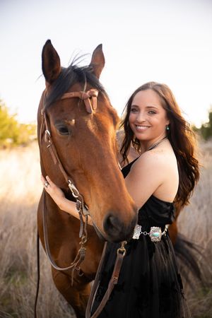 A woman in a black dress poses with a brown horse in a sunlit field at golden hour.