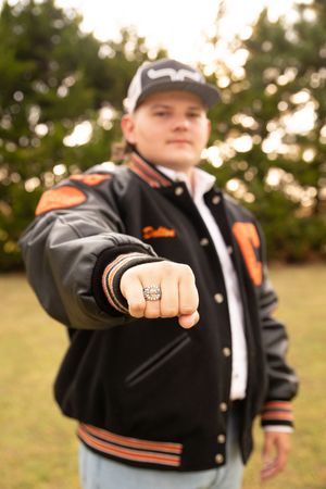 A person in a black varsity jacket with orange trim shows off their championship ring in multiple poses.