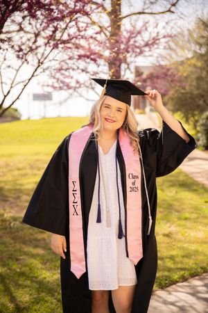 Graduate in black cap and gown with pink stole poses outdoors during spring cherry blossom season.