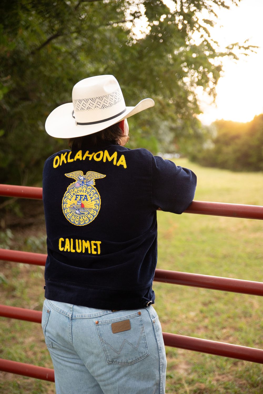 FFA member wearing cowboy hat and blue jeans leans on red metal fence at sunset in Oklahoma.