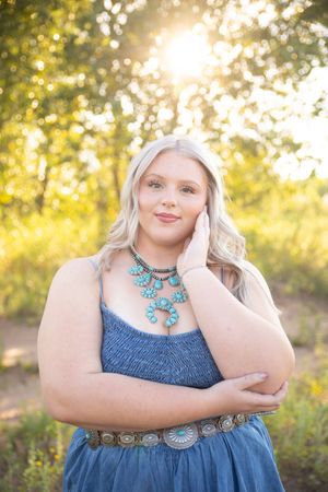 A person in a blue dress and turquoise necklace poses outdoors during golden hour with soft bokeh lighting.