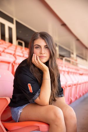 A person in a black shirt sits casually on orange stadium seats while resting their chin on their hand.