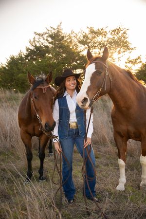 Two brown horses stand in tall grass at sunset with a rider in western attire holding their reins.