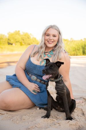 A person in denim sits with a black French Bulldog puppy on sandy ground during golden hour.