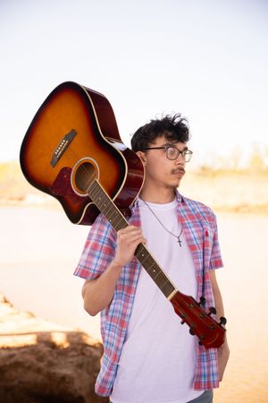 A person in a plaid shirt and white t-shirt poses with an acoustic guitar in a desert landscape at sunset.