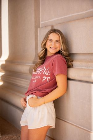 A person in a burgundy t-shirt and white shorts poses casually against a beige wall with natural lighting.