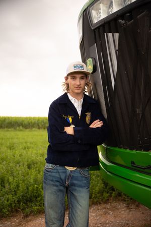 A young person in FFA jacket and baseball cap stands next to a green tractor in a grassy field.