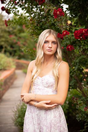 A figure in a floral lace dress poses in a garden setting with vibrant red roses and greenery in the background.