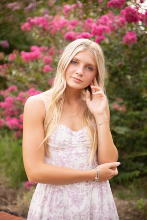 A person in a white floral dress poses in front of vibrant pink flowering bushes in an outdoor garden setting.