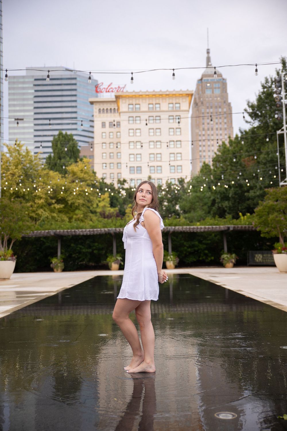 Someone in a white dress stands in a reflecting pool with downtown buildings and skyscrapers in the background.