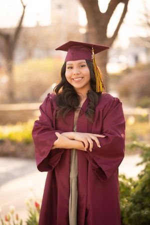 Graduate in maroon cap and gown poses outdoors during golden hour with a warm smile.