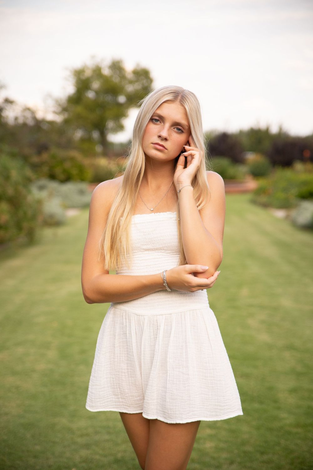A person in a white summer dress stands in a lush garden setting while talking on a phone.