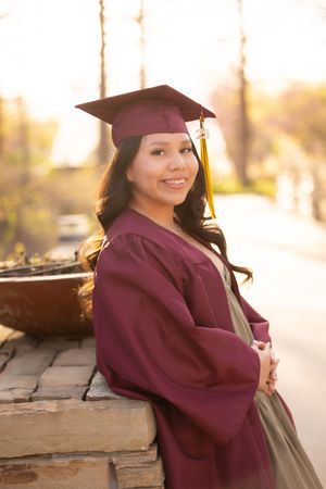 Graduate in burgundy academic regalia poses against stone wall during sunset photography session.