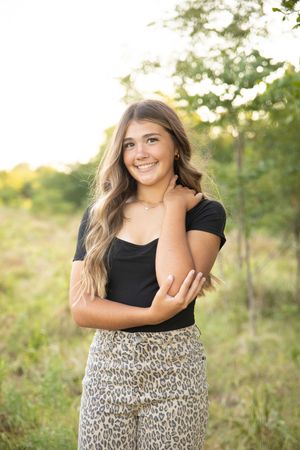 A person in a black top and leopard print pants poses outdoors in a natural setting during golden hour.