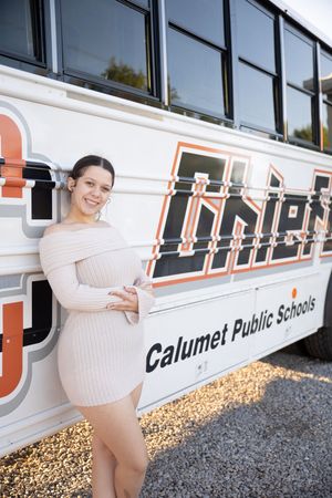 Woman in white sweater dress poses playfully against vintage school bus in outdoor photoshoot.
