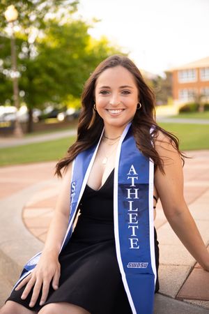 Graduate wearing blue and white stole poses for outdoor graduation photos in black dress on sunny day.