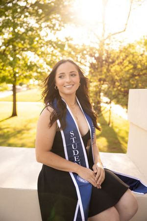 Graduate in navy blue sash and black dress poses outdoors during golden hour sunset for graduation photos.