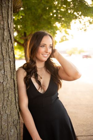 Portrait series of woman in black sleeveless top leaning against tree trunk at sunset with warm natural lighting.