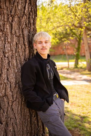 Person in black jacket poses against tree trunk in autumn park setting with golden foliage in background.
