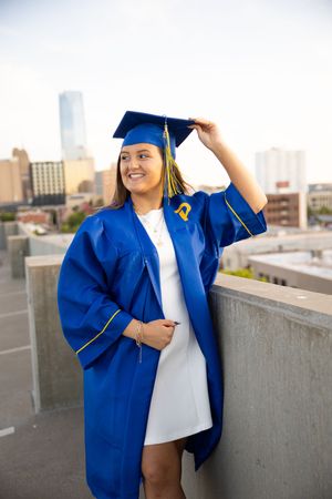 Graduate in blue cap and gown poses on rooftop with city skyline backdrop during graduation photoshoot.
