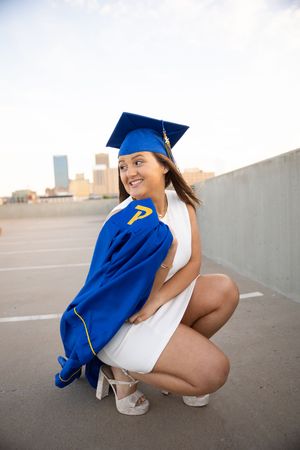 Graduate in blue cap and gown crouches down for a series of creative graduation photos in an outdoor setting.