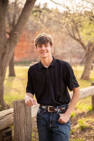 Casual portrait of someone in a black polo shirt leaning on a wooden fence in an autumn outdoor setting.