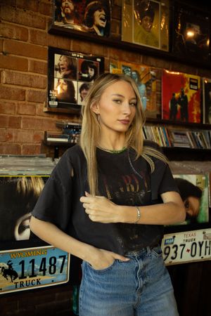 A person in a black t-shirt and jeans leans casually against a brick wall in a dimly lit room with license plates visible.