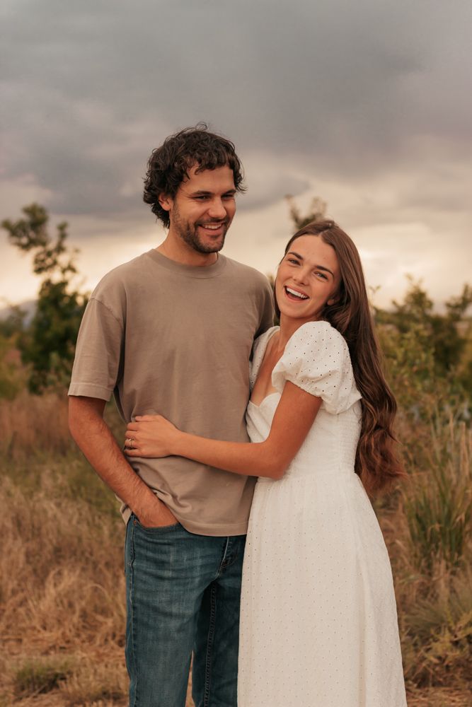 A couple embraces and laughs together in a field during golden hour with stormy clouds | Westminster, CO motherhood & family photographer