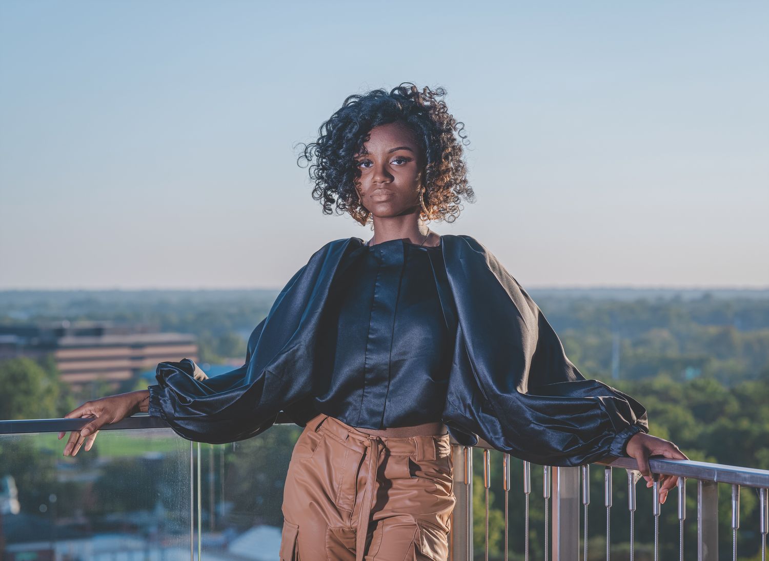 An African-American high school female in a black fashionable top and brown, leather pants poses on a balcony overlooking the city.