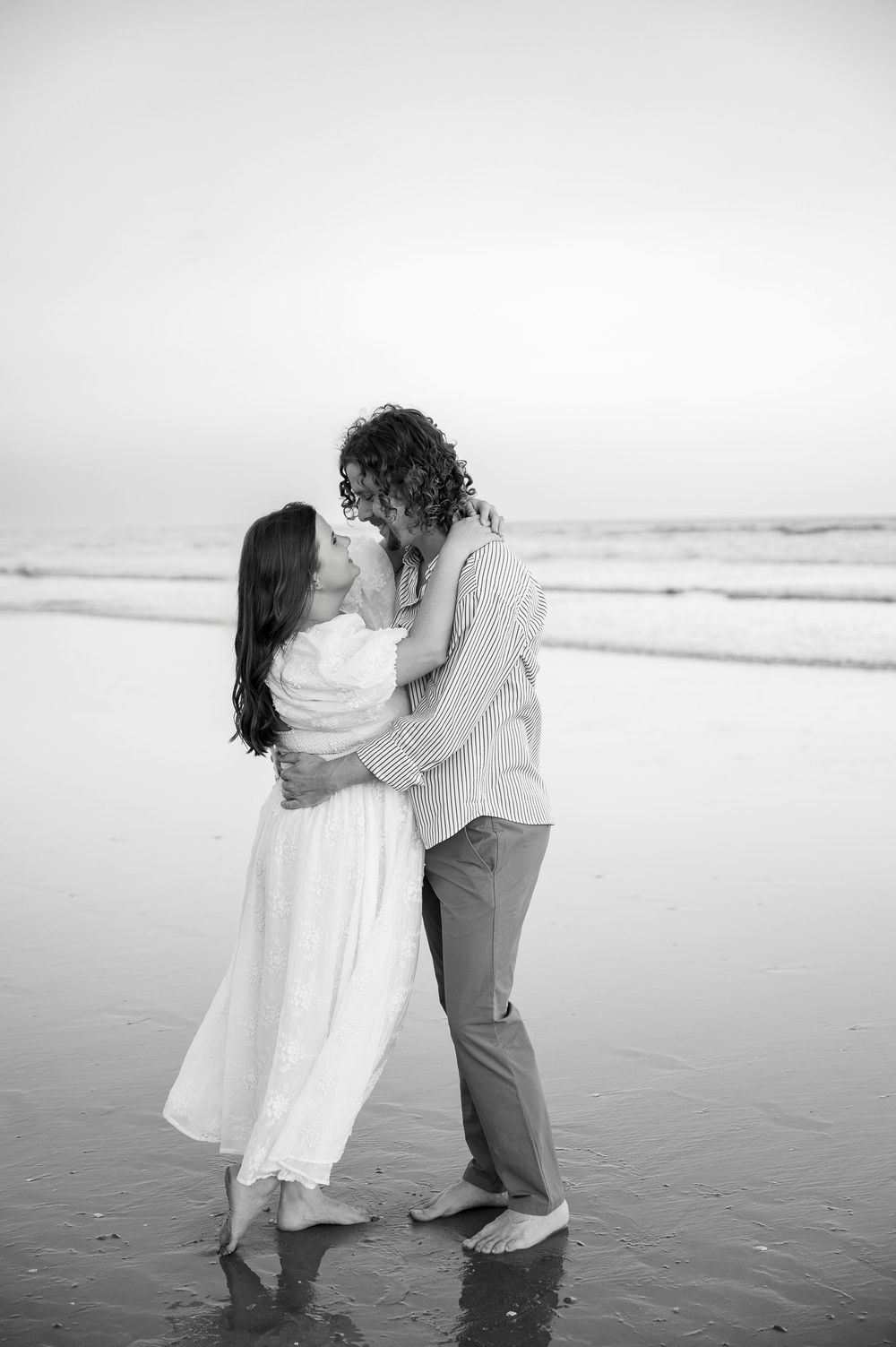 A black and white photo captures a romantic dance on the beach with waves in the background.