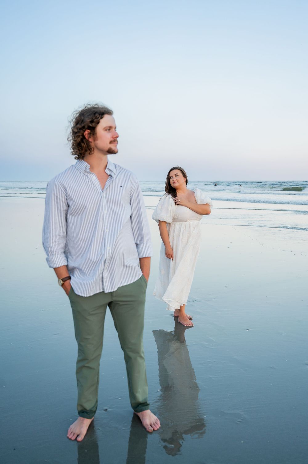 Two people walk barefoot along the wet sand of a beach during sunset.