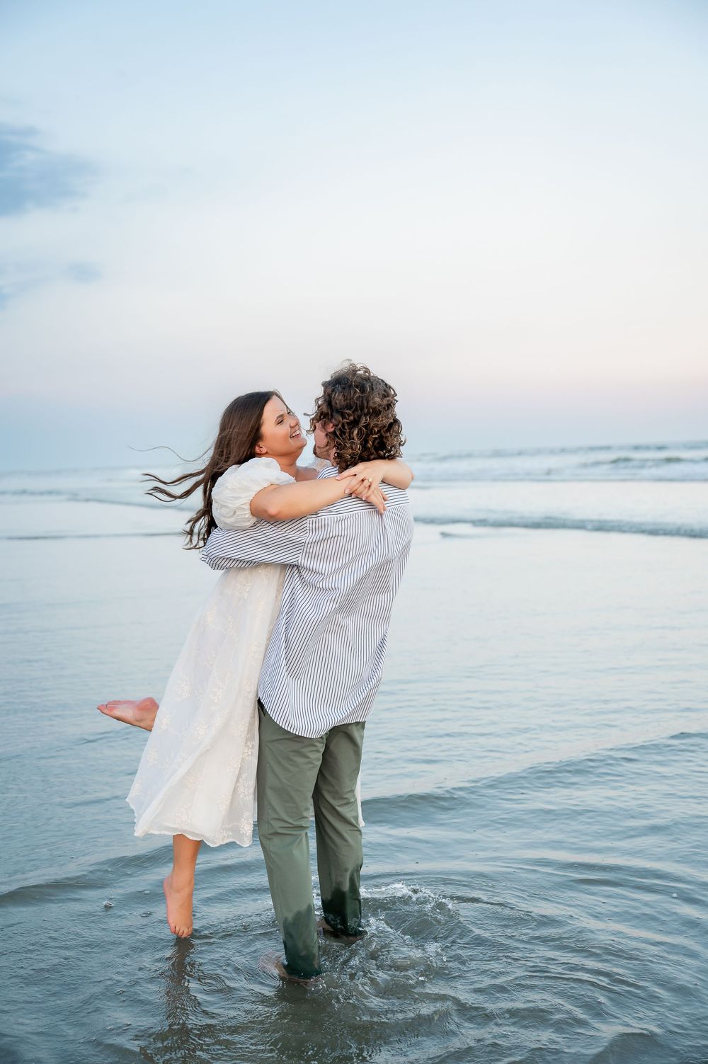 A playful couple splashes in shallow ocean water while wearing white and grey casual attire.