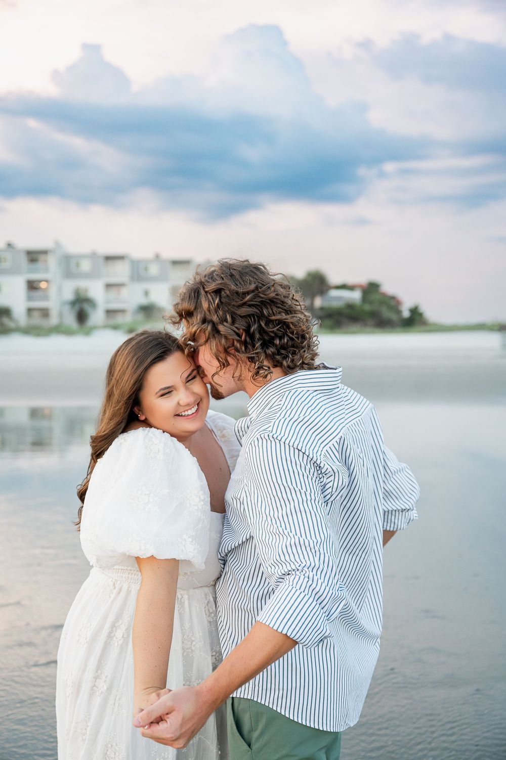 A couple shares an intimate moment while standing in shallow beach water at dusk.