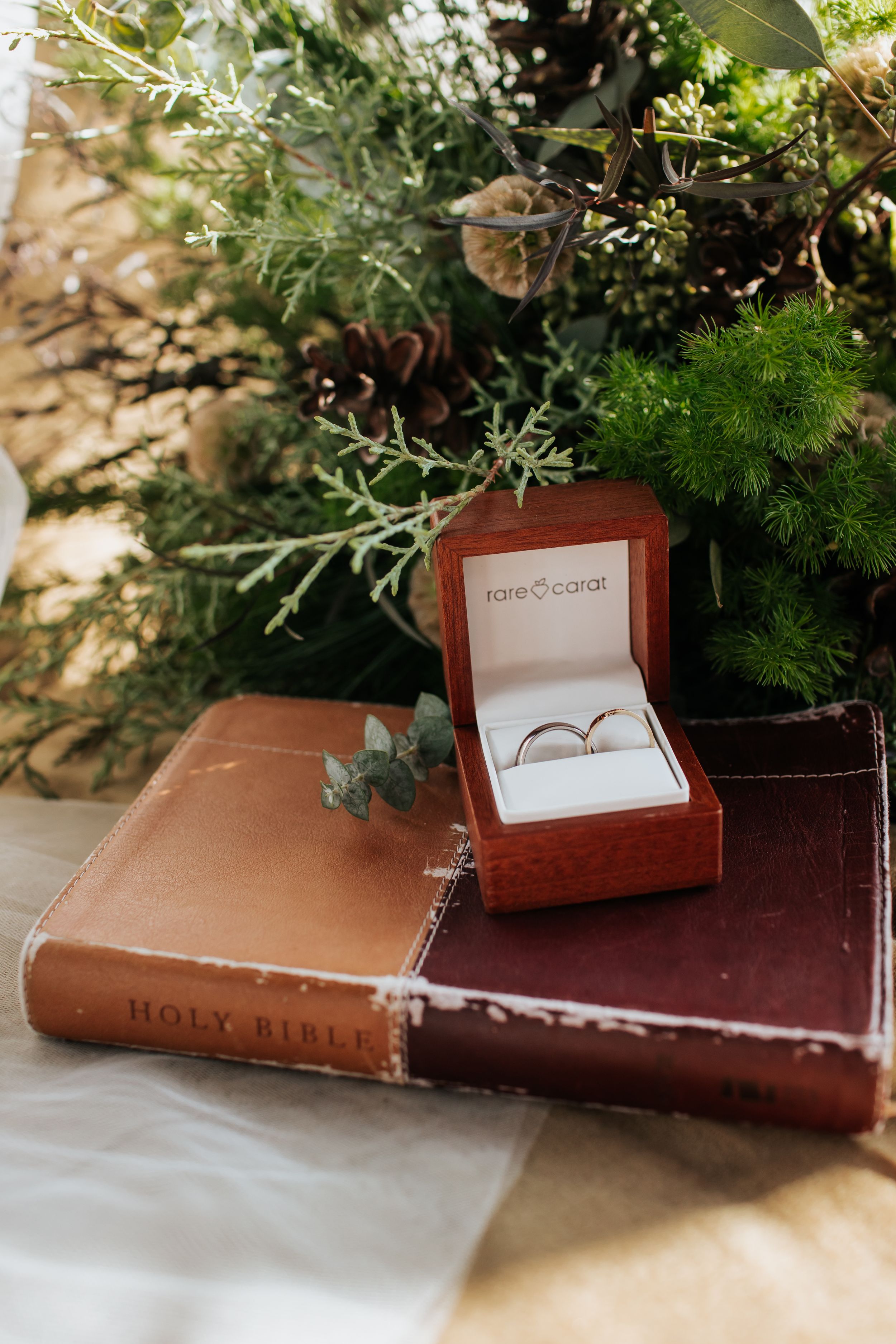 A Holy Bible lies open with a brown wedding ring box nestled among green foliage decorations.