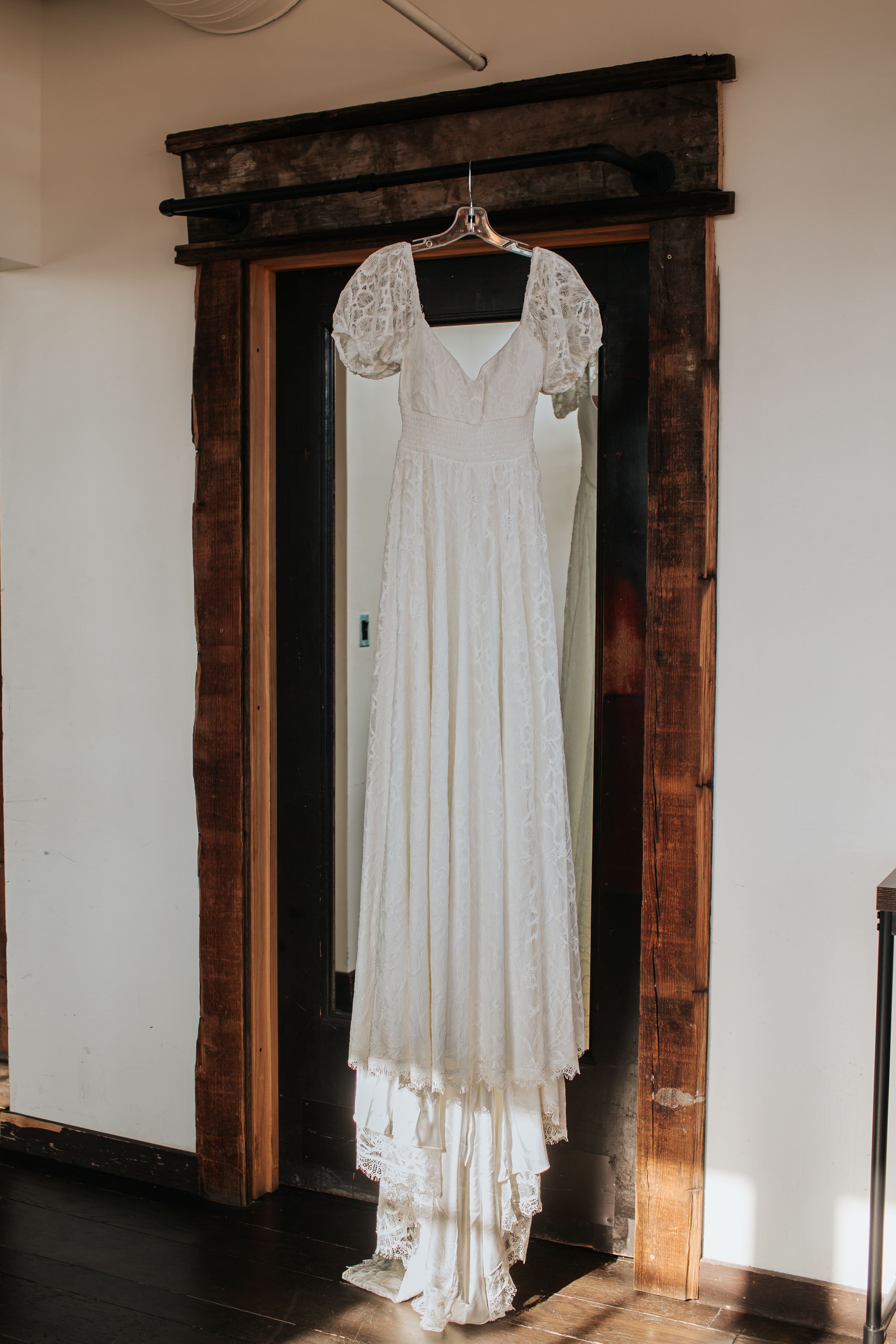 White lace wedding dress hangs elegantly against rustic wooden door frame in bridal preparation room.