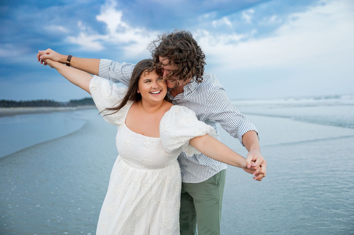 Two people share a playful moment while standing barefoot on a beach with blue skies overhead.