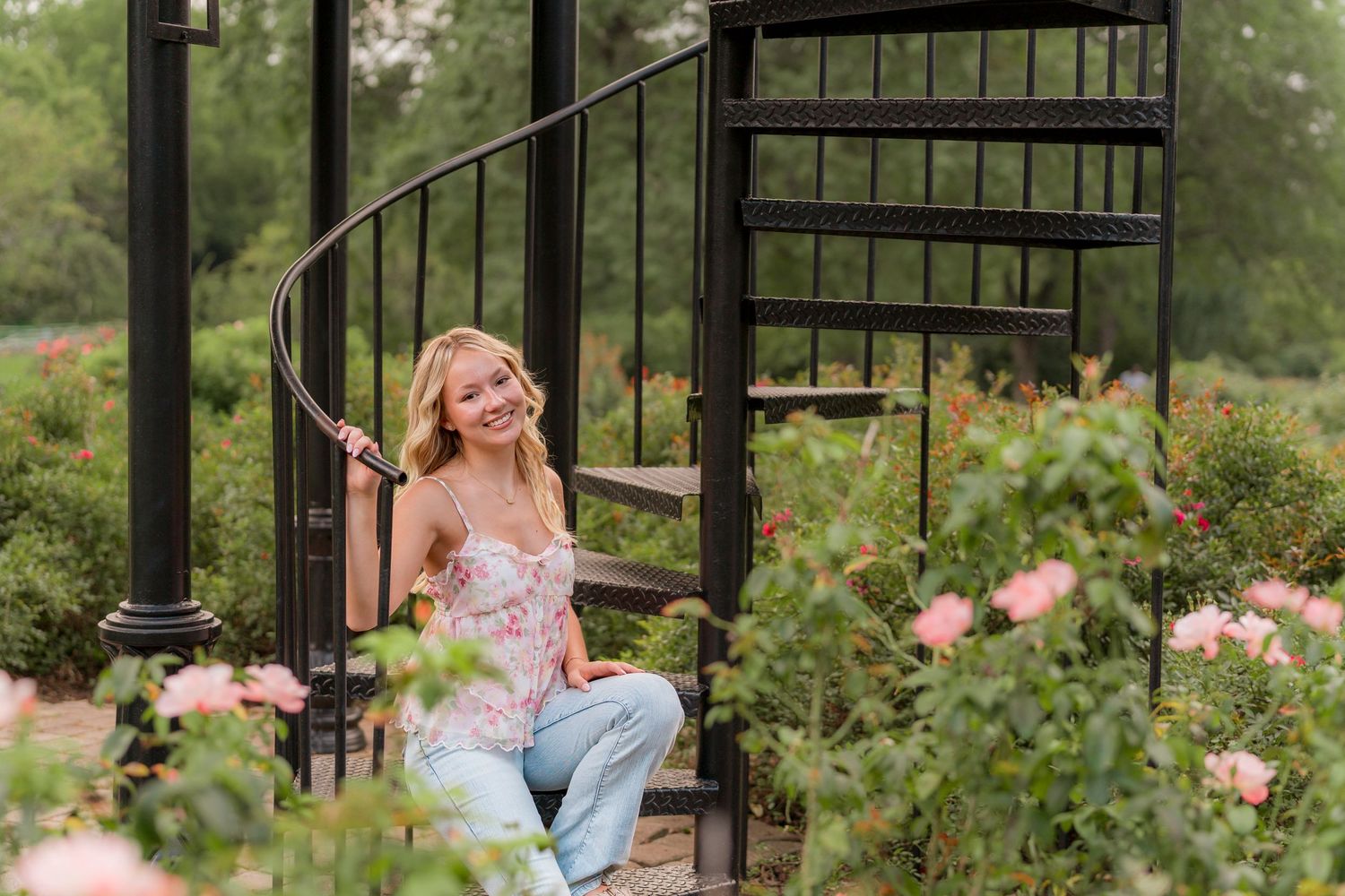 Someone sits on black spiral staircase surrounded by blooming pink roses in a garden setting.