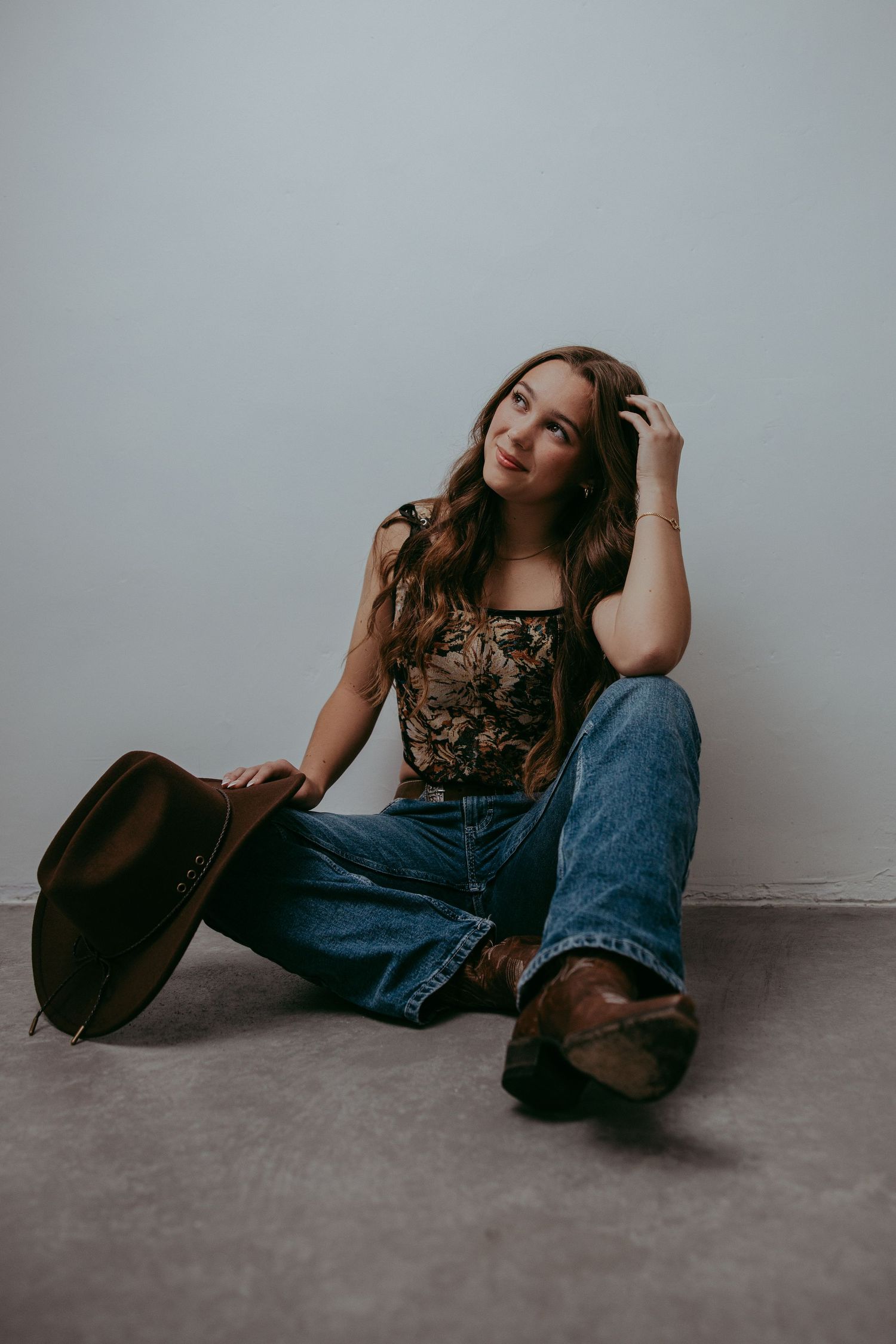A person in a floral top and jeans sits casually on the floor holding a brown hat against a gray background.