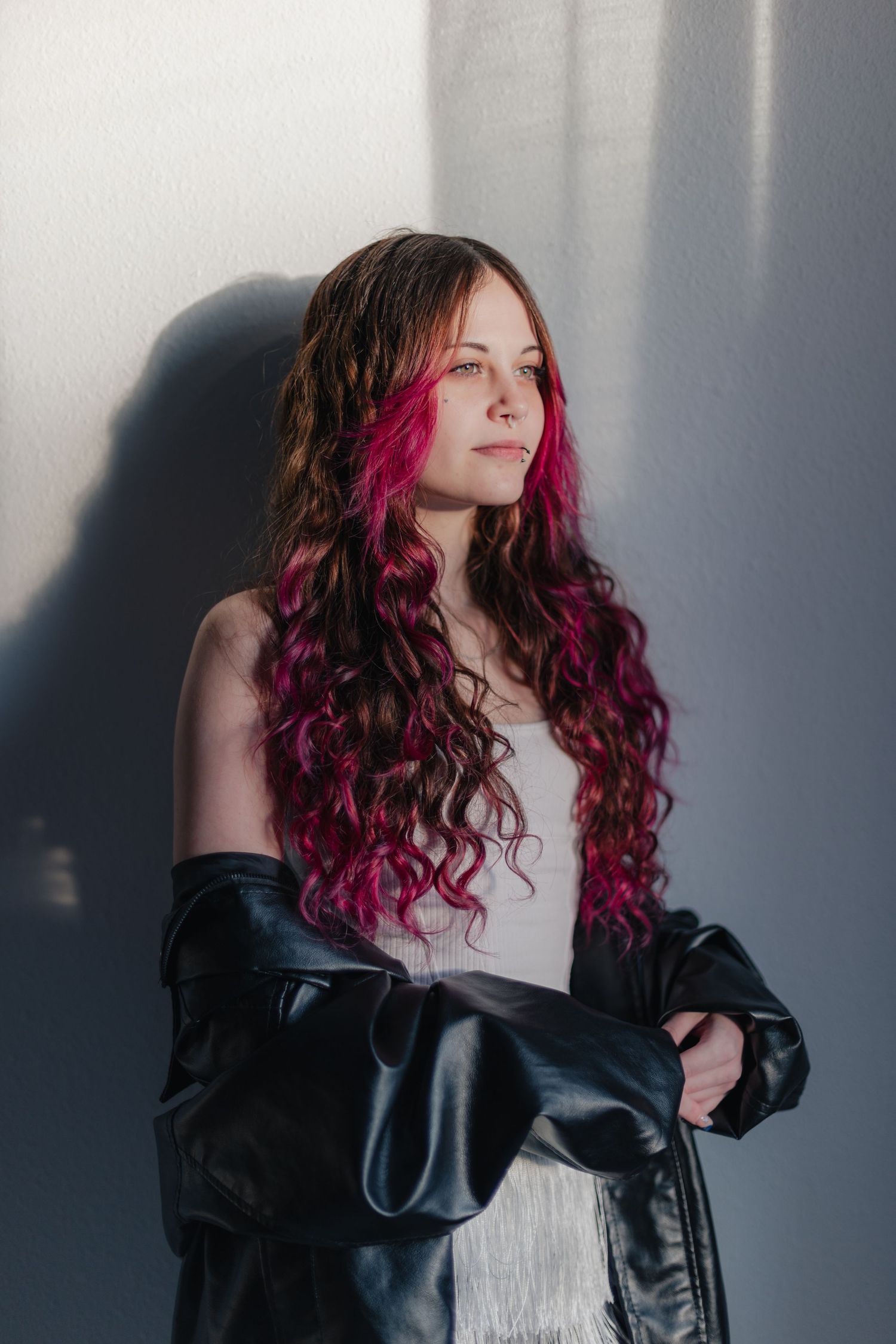 A person with vibrant magenta curly hair poses against a gray wall wearing a black leather jacket and white top.