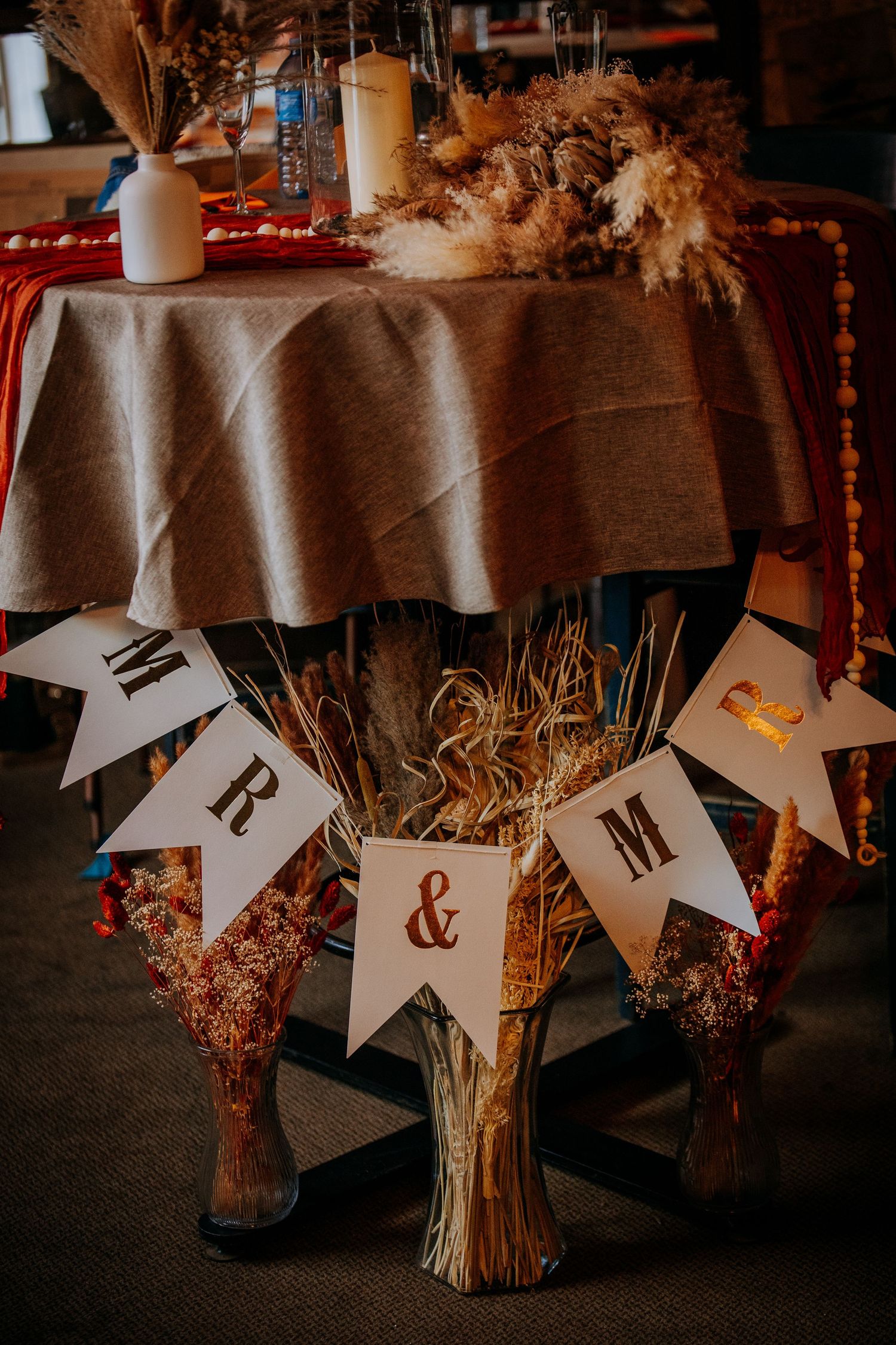 Rustic wedding decor featuring a Mr & Mrs banner with dried flowers and moody lighting at a reception table.