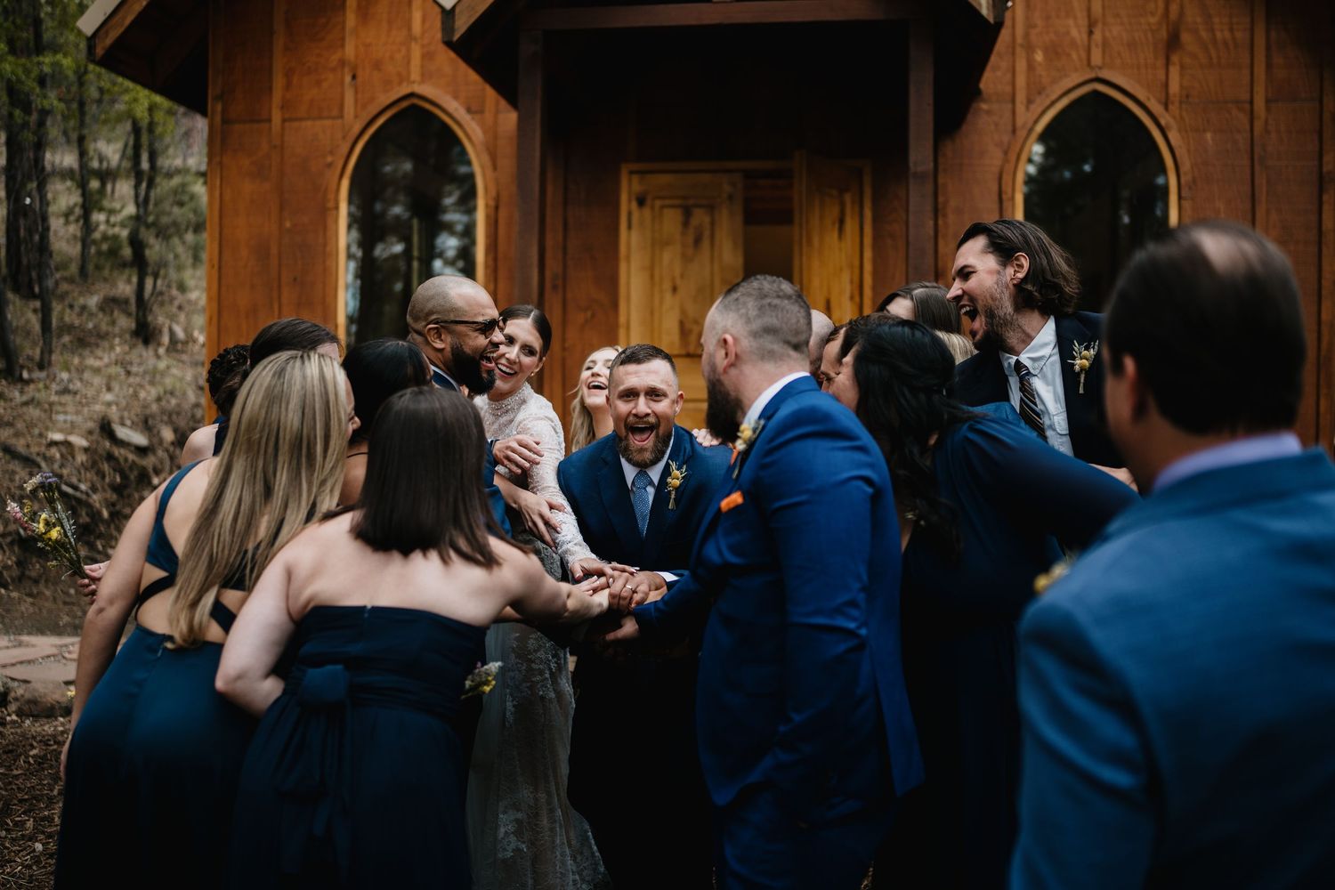 Wedding party celebrating outside rustic wooden chapel with bridesmaids in navy dresses and groomsmen in blue suits at Flying J Ranch